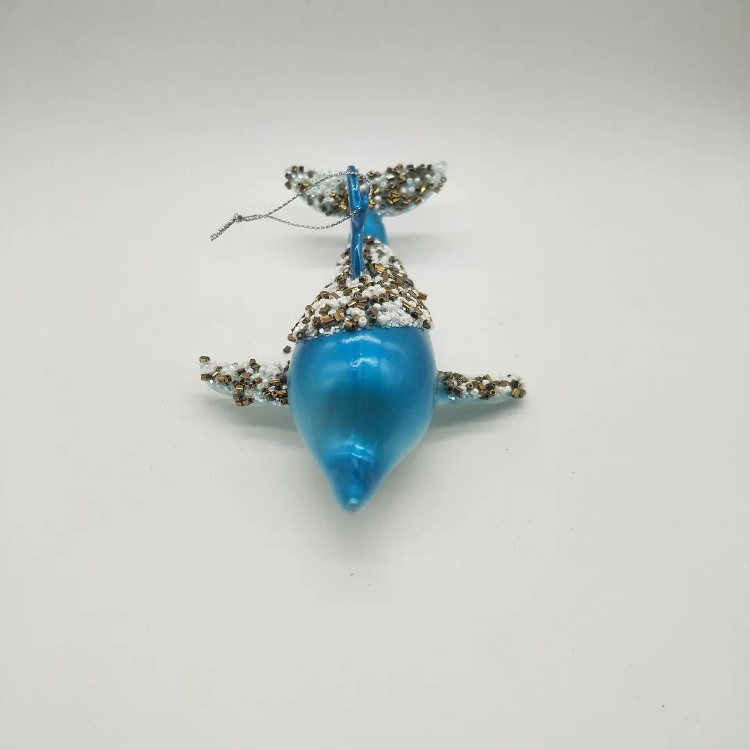 This product is a glass Christmas ornament in the shape of a whale. It is decorated with blue and gold paint and has a sparkling finish.
